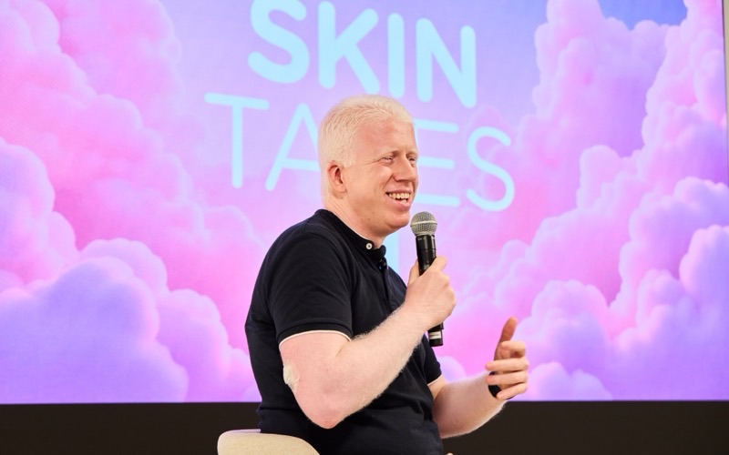 Rubén, con un micrófono y un polo negro, habla sonriendo sentado delante de una pantalla con el logotipo de Skin Tales sobre nubes rosas.