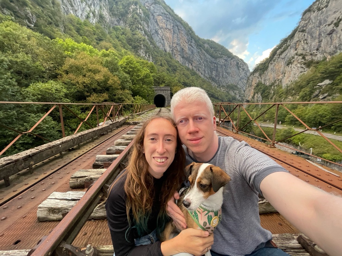 Rubén, su pareja y su perrita, abrazados en un selfi sobre un viejo puente de ferrocarril, con un túnel y montañas detrás.